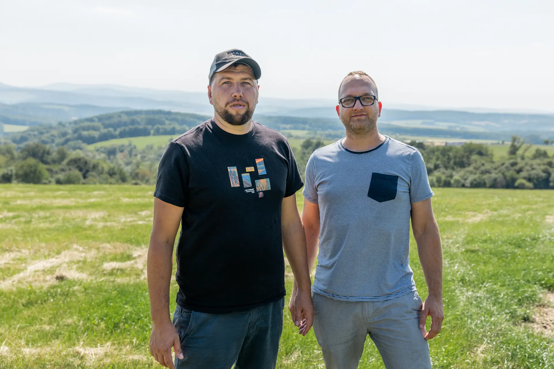 Two men standing in a field with a landscape in the background.