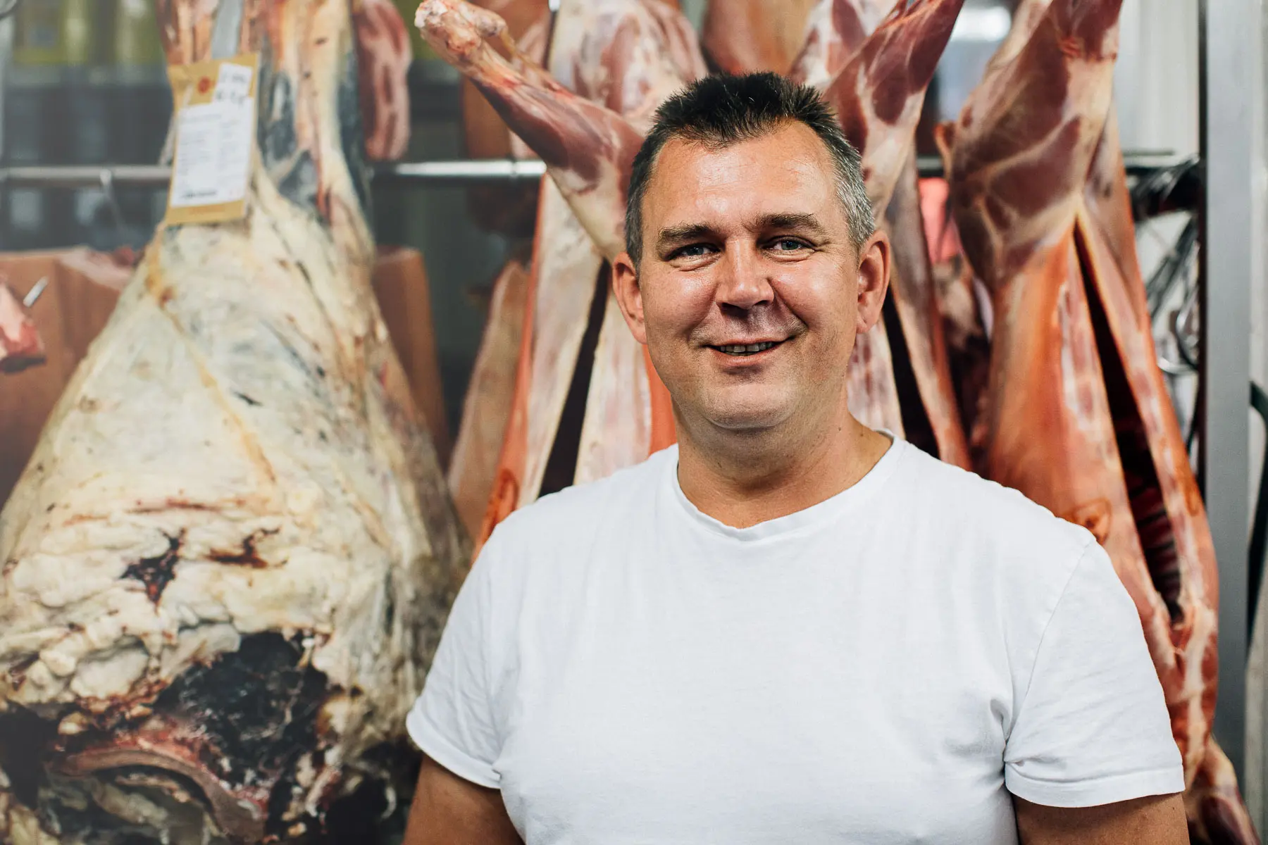 Butcher in a white T-shirt standing in front of hanging beef carcasses.